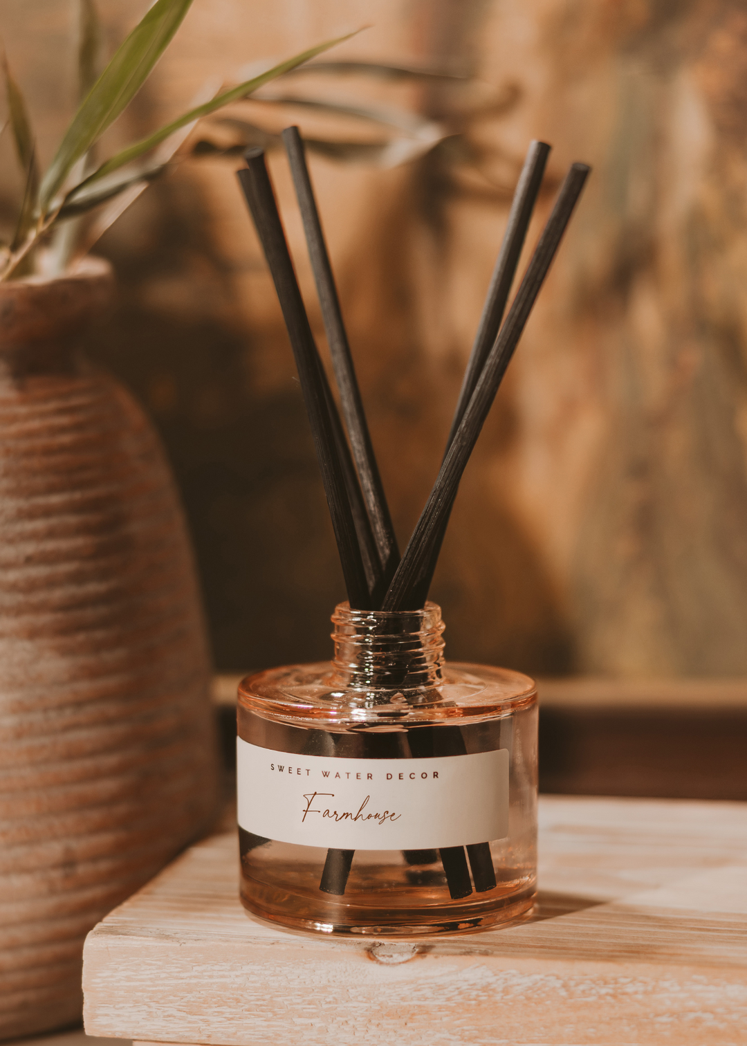 Diffuser with reeds on a wooden surface next to a plant. Farmhouse Diffuser by Sweet Water Decor.