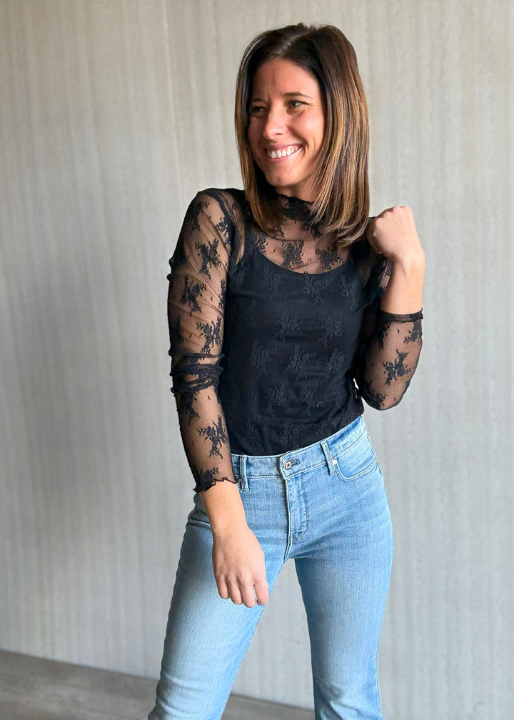 Woman wearing a black lace top and light blue jeans against a neutral background. Women's Long-Sleeve Black Lace Sheer Layering Top.