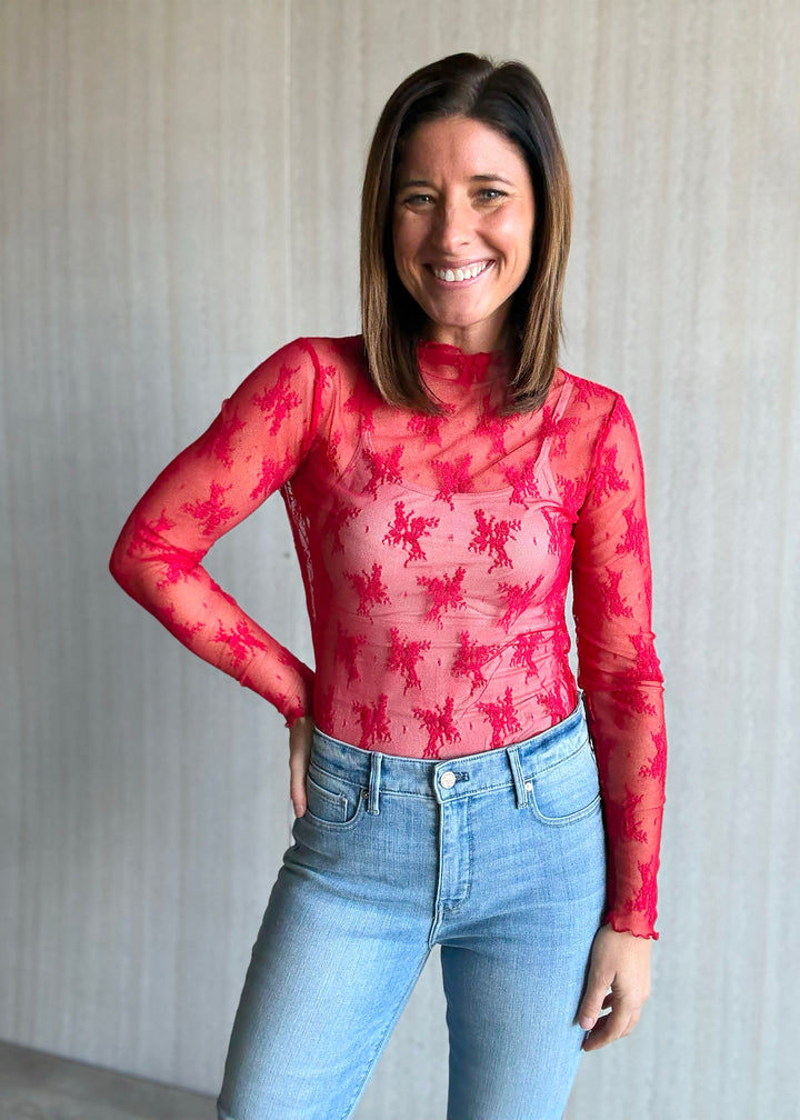 Woman wearing a red floral top and blue jeans against a neutral background. Sheer Red Lace Long-Sleeve Layering Top.