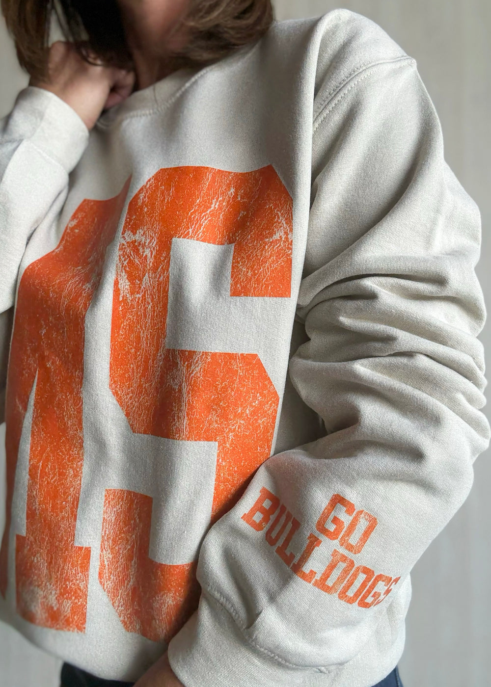 Person wearing a white sweatshirt with orange text and graphics | Women's Mahomet Seymour Cream and Orange Sweatshirt Fanwear