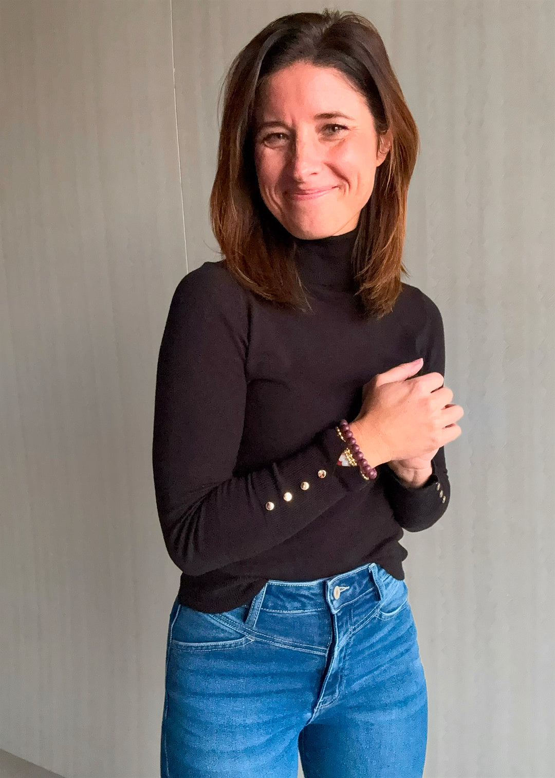 Woman wearing a black turtleneck with gold embellishments and blue jeans against a neutral background