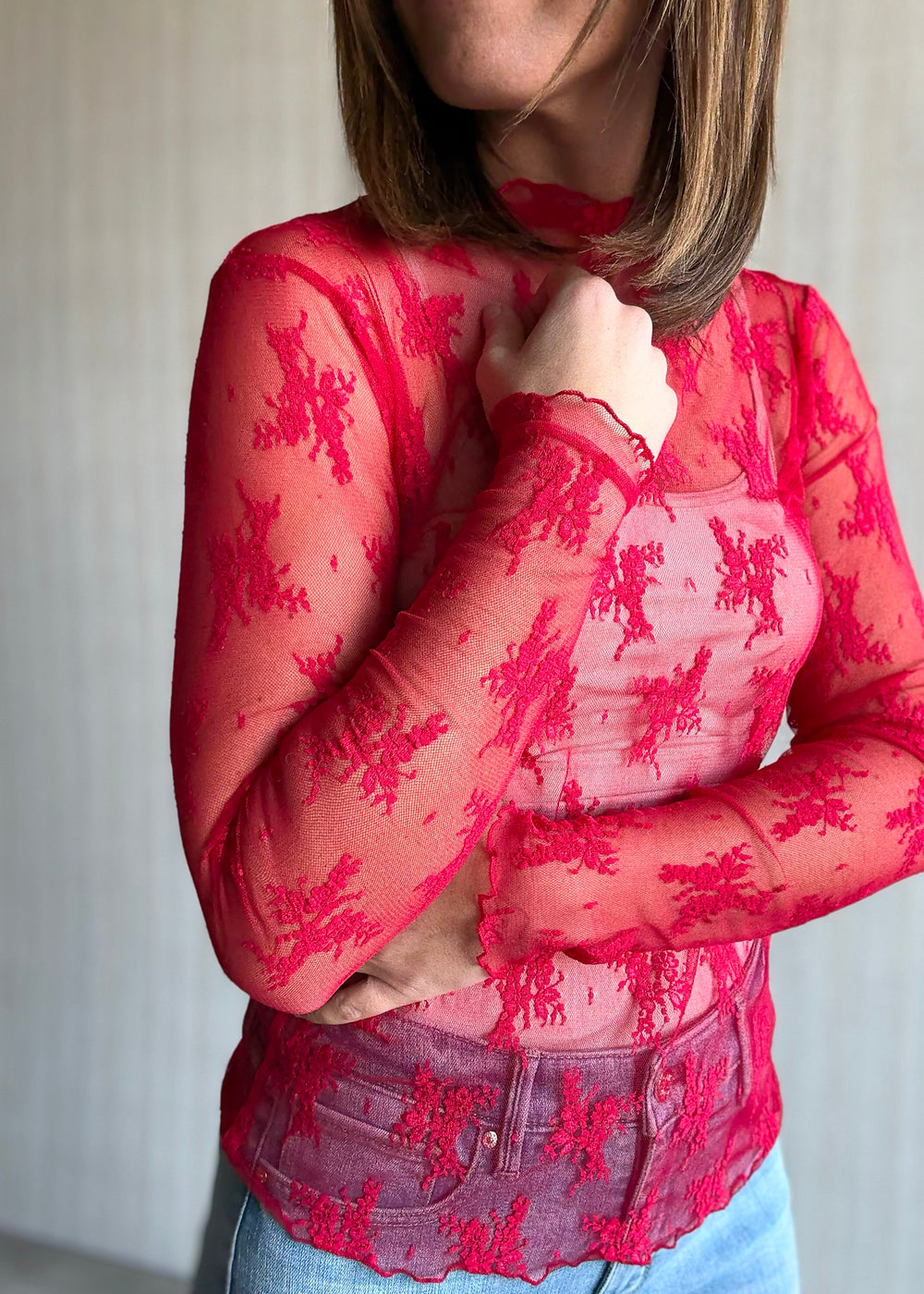Woman wearing a red floral top and blue jeans against a neutral background. Sheer Red Lace Long-Sleeve Layering Top.