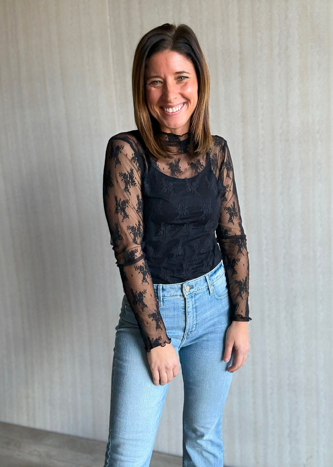 Woman wearing a black lace top and light blue jeans against a neutral background. Women's Long-Sleeve Black Lace Sheer Layering Top.