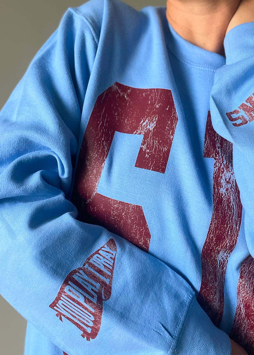 SJO Spartans Sweatshirt - Columbia blue crewneck sweatshirt with "SJO" in distressed maroon letters prominently displayed on the front. Each sleeve has maroon text at the sleeve with one stating "GO SPARTANS" and the other with the “You Play I Pray”