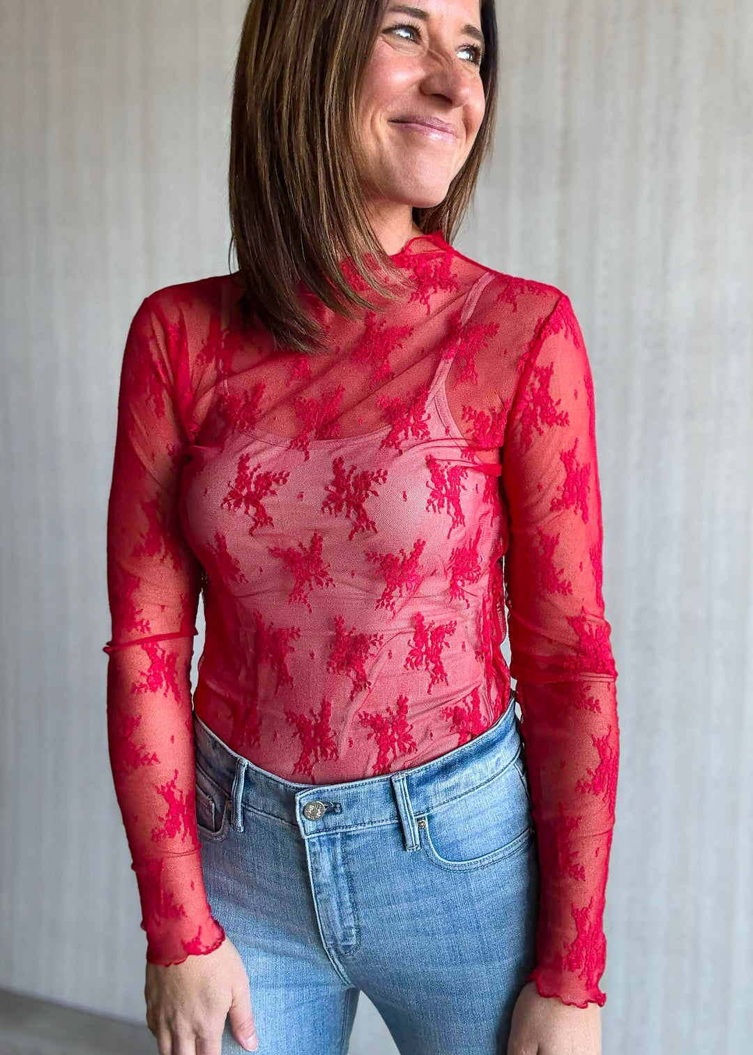 Woman wearing a red floral top and blue jeans against a neutral background. Sheer Red Lace Long-Sleeve Layering Top.