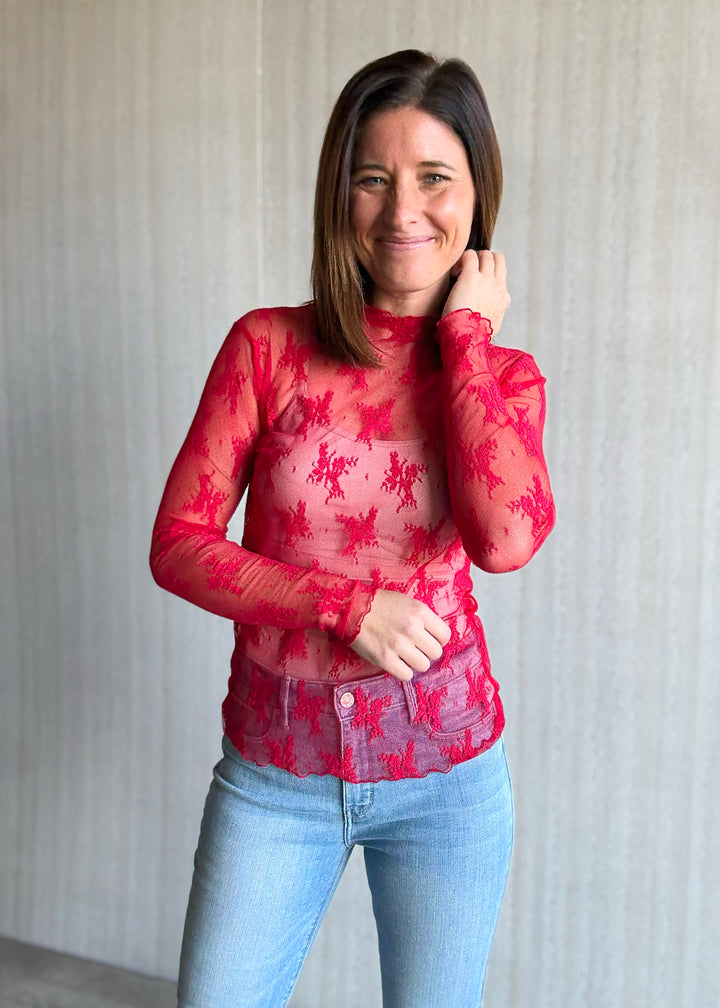 Woman wearing a red floral top and blue jeans against a neutral background. Sheer Red Lace Long-Sleeve Layering Top.
