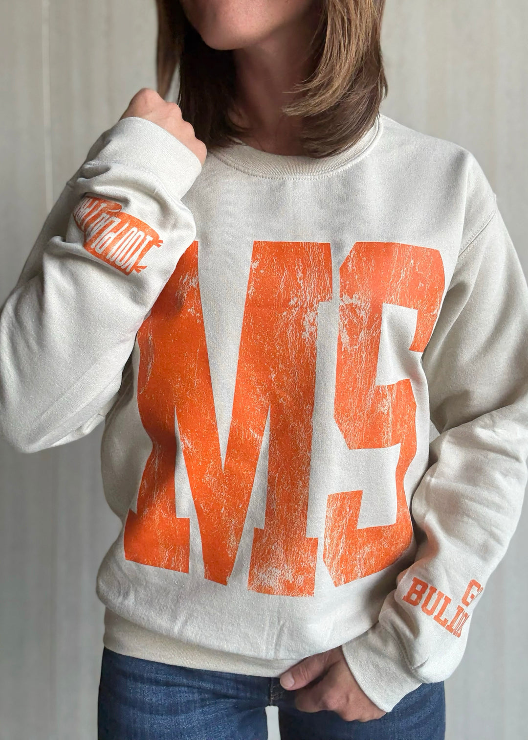 Person wearing a white sweatshirt with orange text and graphics | Women's Mahomet Seymour Cream and Orange Sweatshirt Fanwear