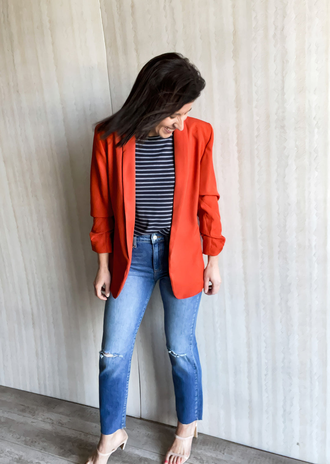 Orange Blazer with ruched sleeves paired over a navy and white tank top and knee slit jeans.