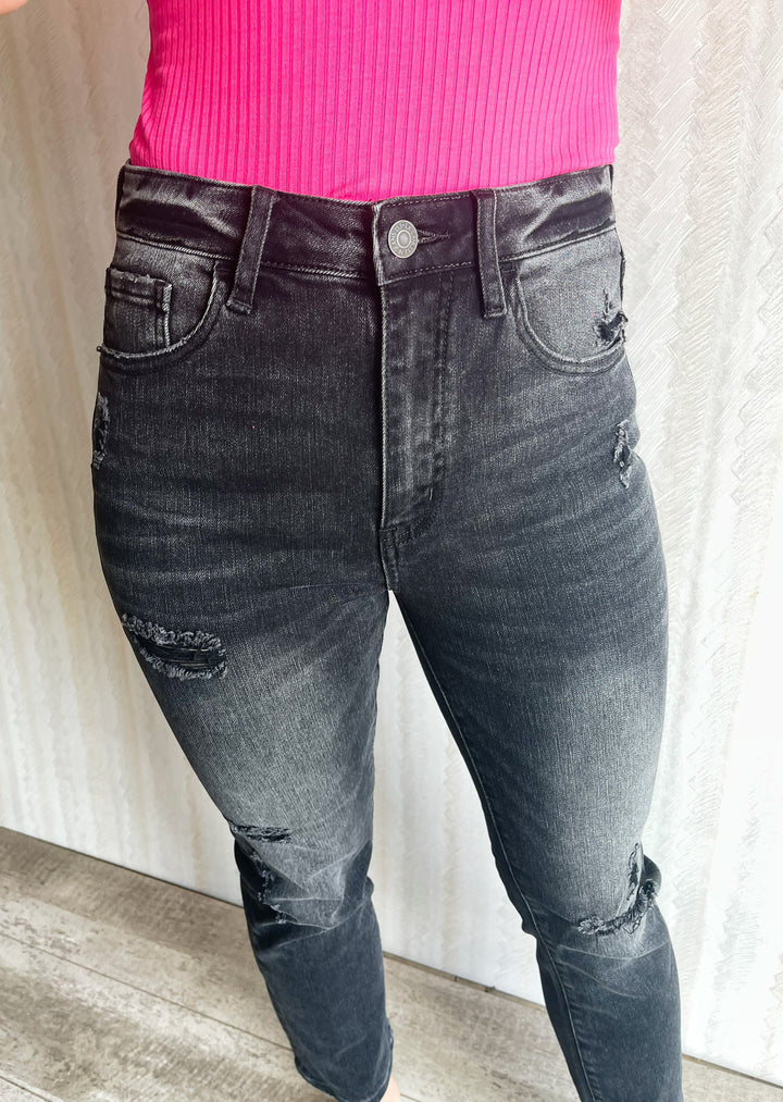 Risen Straight leg jeans in a vintage black wash with slight fading on the front legs staring on the upper hip down to the knee as well as on the back bottom and behind the knees. These have distressing throughout, including a higher distressed ankle on the back of each leg. Paired with a pink tank top. 